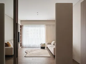 A modern living room featuring neutral-colored furniture, a patterned rug, and large windows with sheer curtains.