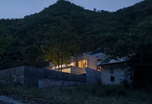 一座擁有混凝土外牆的現代住宅，窗戶透出燈光，四周環繞著樹木。