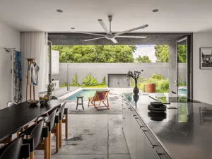 A modern kitchen with dark concrete countertops, wooden cabinets, and a glass backsplash, featuring a large table with chairs