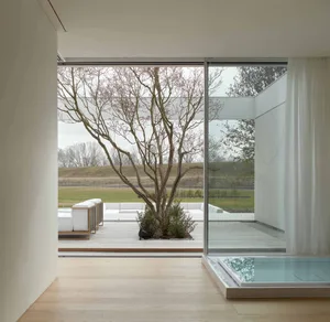 A modern living room with a large glass window overlooking an outdoor garden with a bare tree and white furniture.