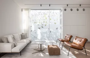 A modern residential interior featuring a white sofa with brown leather accents, a glass-top coffee table, and a woven ottoma