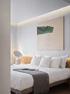 A minimalist bedroom with white bedding, a grey throw blanket, and a peach-colored abstract painting hanging above the bed. T