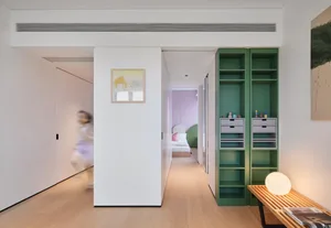 A modern apartment interior featuring a hallway with light wood floors, green shelving, a bedroom with a pink wall, and a lar