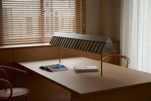 A minimalist, metal desk lamp is placed on a wooden table in a room with wooden blinds and a white curtain.