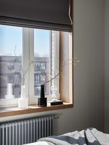 A minimalist bedroom with a wooden window sill decorated with vases and branches, featuring a radiator and a carpet on the fl