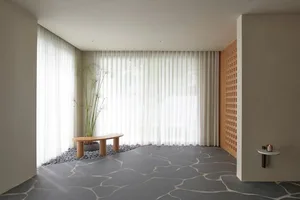 A minimalistic living room featuring a grey, stone-patterned floor, a white sheer curtain, and a wooden bench seat with a pla