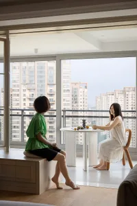 Two women sitting in a modern living room with white tiles, looking out of a large window with a cityscape view.
