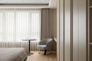 A minimalist bedroom featuring taupe textiles and light hardwood flooring, with sheer curtains allowing soft light to filter 