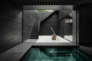 Modern minimalist interior space featuring concrete walls and floor, with a central rock sculpture and a glass staircase.