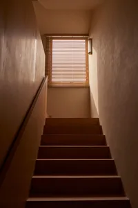 A modern staircase with wooden steps illuminated by natural light coming through a window with wooden blinds.