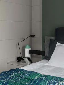 A minimalist bedroom with concrete, metal, and fabric materials, featuring a bed with a green patterned blanket, a modern lam