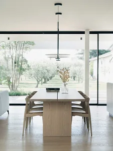 Modern minimalist dining room featuring a wooden table and chairs, with floor-to-ceiling windows showcasing an outdoor olive 
