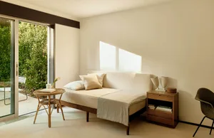 A minimalist bedroom featuring a cream-colored daybed with white and cream bedding, a small wooden side table with books and 