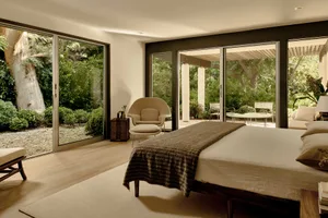 A minimalist bedroom with a bed, chair, and large windows overlooking greenery.