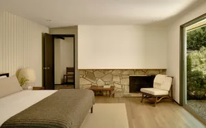 A minimalist bedroom featuring a bed with a wooden headboard, a stone fireplace, and a lounge chair near a large window.
