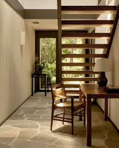 A modern home office with wooden furniture and stone flooring, featuring a built-in desk, chair, and large window.