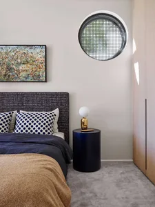 A minimalist bedroom with a round window, fabric headboard, and glass bedside table.