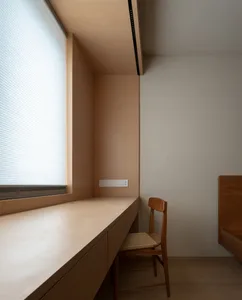 A minimalist home office features a wooden counter and chair against a white wall, with a window allowing natural light in.