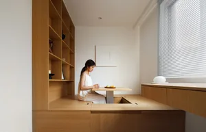 A woman sits at a small table with a fruit bowl in a modern interior design setting with wooden elements and soft lighting.