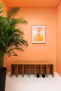 A minimalist modern entryway with a wooden shoe shelf against an orange wall, featuring two pairs of black sandals at the bas