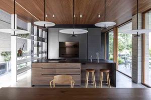 Modern kitchen with wood finishes and a minimalist design, featuring an island with stools and a view of the surrounding spac