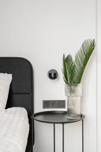 Modern minimalist bedroom featuring a black metal bedside table with a glass vase containing palm leaves.
