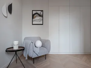 A minimalist bedroom featuring a grey upholstered chair, a small side table, and a framed abstract art piece.
