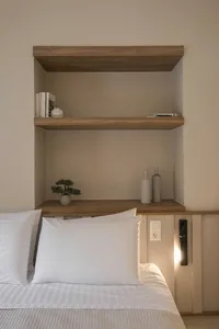 A minimalist bedroom featuring a bed with white bedding, a wooden headboard, and concrete shelves with books and decorative i