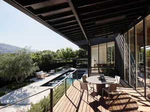 Modern residential deck featuring wood flooring, glass walls, and a pool with surrounding greenery.