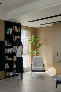 A minimalist living room featuring a large bookshelf, a single armchair, and a potted plant in front of a window.