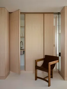A minimalist bedroom with wooden walls and floor, featuring a chair in the foreground and a small kitchen area in the backgro