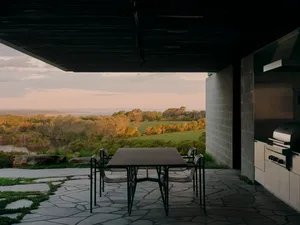 A minimalist outdoor dining area featuring concrete and stone materials, with a table and chairs overlooking a scenic landsca