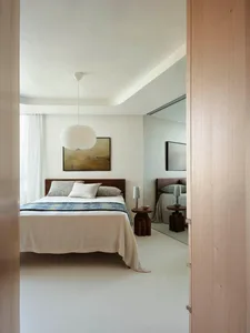 A modern bedroom with neutral tones featuring a bed with white bedding, a hanging white light fixture, and minimalist decor.