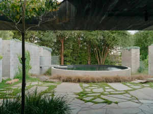 A serene outdoor space featuring a round pool surrounded by stone walls, surrounded by lush greenery and a pathway of irregul