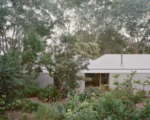 Modern house exterior with white concrete walls and a gray corrugated metal roof, surrounded by lush greenery and trees.
