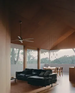 A modern living room with a black leather sofa, wooden ceiling, and large windows overlooking trees.