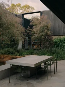 A modern outdoor space featuring a concrete table and chairs surrounded by greenery and a dark building.
