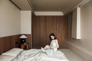 A woman sitting on a bed, petting a cat, with a bedside lamp.