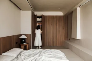 Minimalist bedroom featuring wood paneling, a neatly made bed, and a person choosing clothing from an open closet.