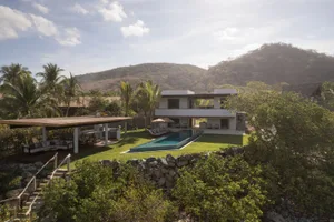 A modern two-story house with a concrete and glass exterior surrounded by lush greenery and a swimming pool.