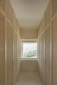 A narrow, light wood corridor with transparent partitions leading to a window view of a green outdoor space.