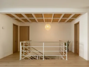 A minimalist interior with white wood flooring and a white wall, featuring a white staircase and wooden ceiling beams.