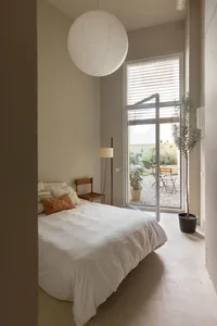 A minimalist bedroom with a white bed, white walls, and a large window for natural light.