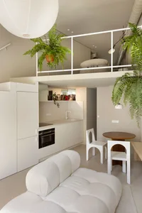 A modern minimalist living room with white furniture, neutral-toned walls, and a small kitchen area.
