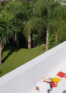 A man with a hat and white shirt stands next to a white wall, examining a piece of paper with samples in a landscaped area wi