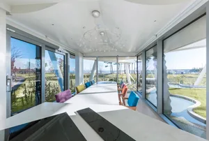 A modern dining room with a long table surrounded by colorful chairs, featuring large glass windows overlooking a green lands
