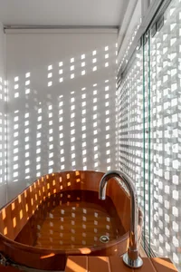 A modern minimalist bathroom with a wooden bathtub under a grid-patterned window, casting sunlit and shadowed square patterns