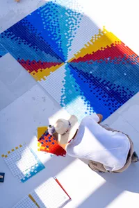 A person installing a colorful mosaic tile floor featuring various shades of blue, white, red, and yellow.