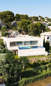 Modern white villa with glass walls and stone accents, surrounded by lush greenery.