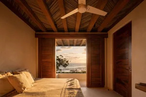 A cozy bedroom featuring wooden beams on the ceiling, a bed with fabric bedding, and a coastal view through the window.
